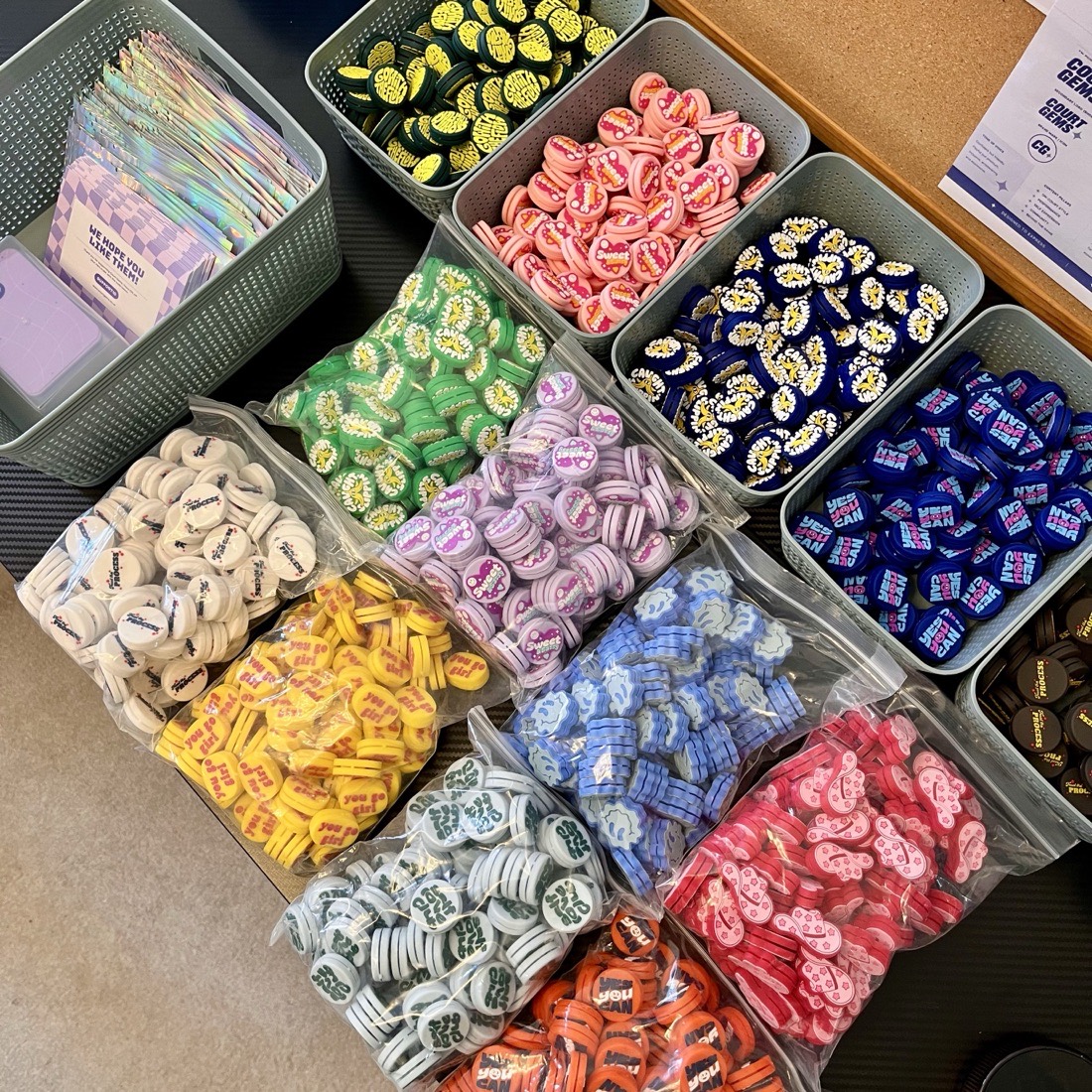 Bins and bags of Court Gems dampeners sorted by design, next to a box of holographic pouches