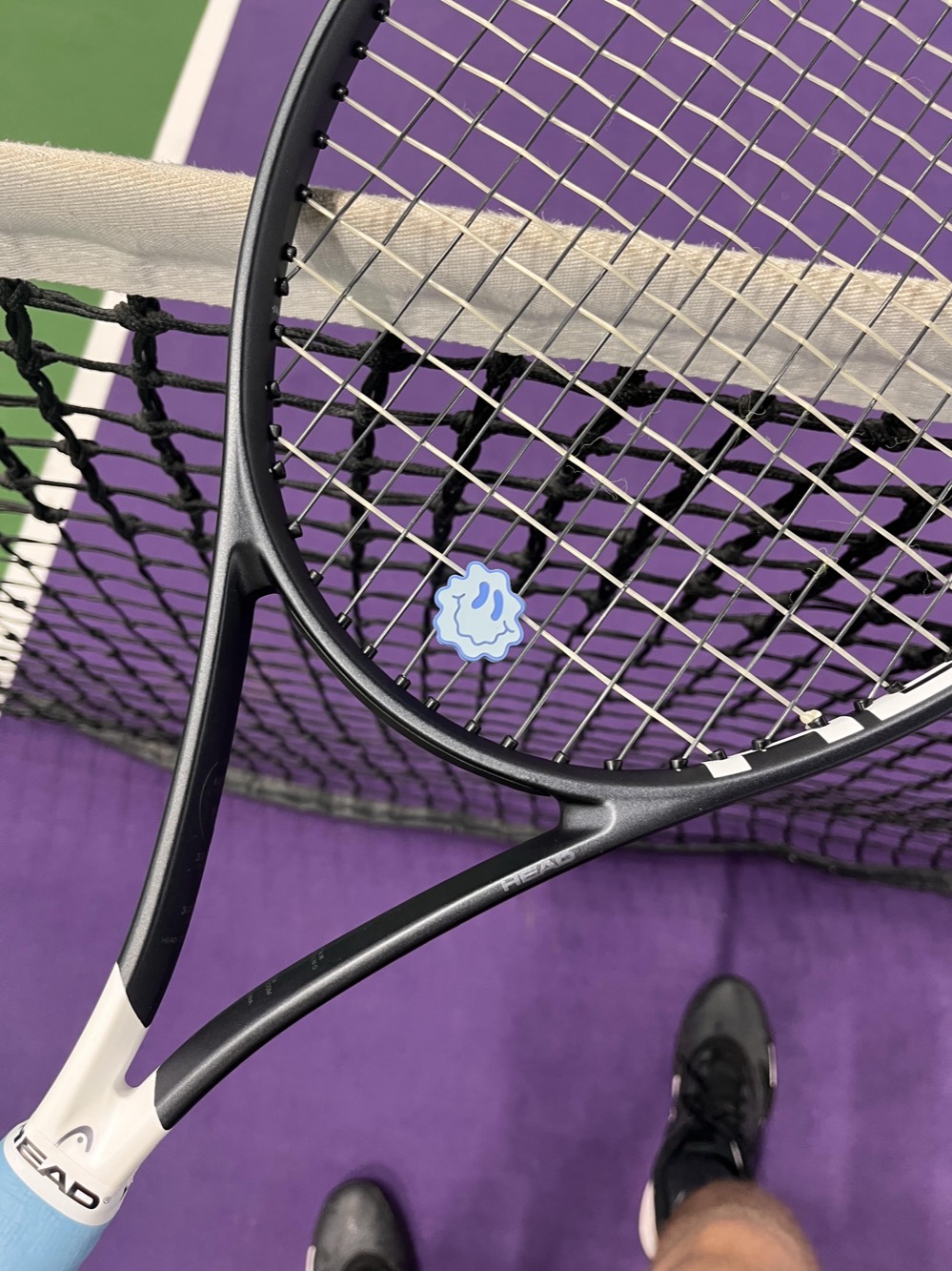 A Smiley Days dampener fitted on a black Head racket, held over the net on an indoor court